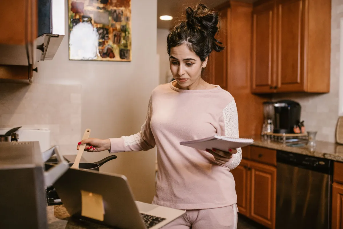 A person standing at a kitchen counter reading a tablet beside an open laptop