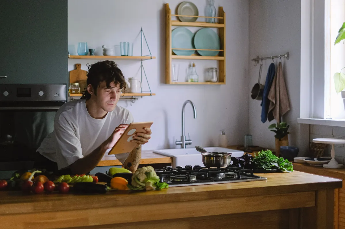 A person leaning on a kitchen island reading from a tablet, with vegetables laid out on the counter